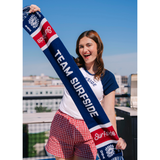 Girl holding Team Surfside USA 250 themed scarf. The Soccer scarf features Team Surfside in the middle, the surfside logo and the sun logo with fringe. The soccer scarf is red, white and blue.