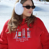 Person wearing a red sweatshirt with 'Surfside Ski Club' design, white ear muffs, and sunglasses in a snowy background.