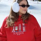 Person wearing a red sweatshirt with 'Surfside Ski Club' design, white ear muffs, and sunglasses in a snowy background.