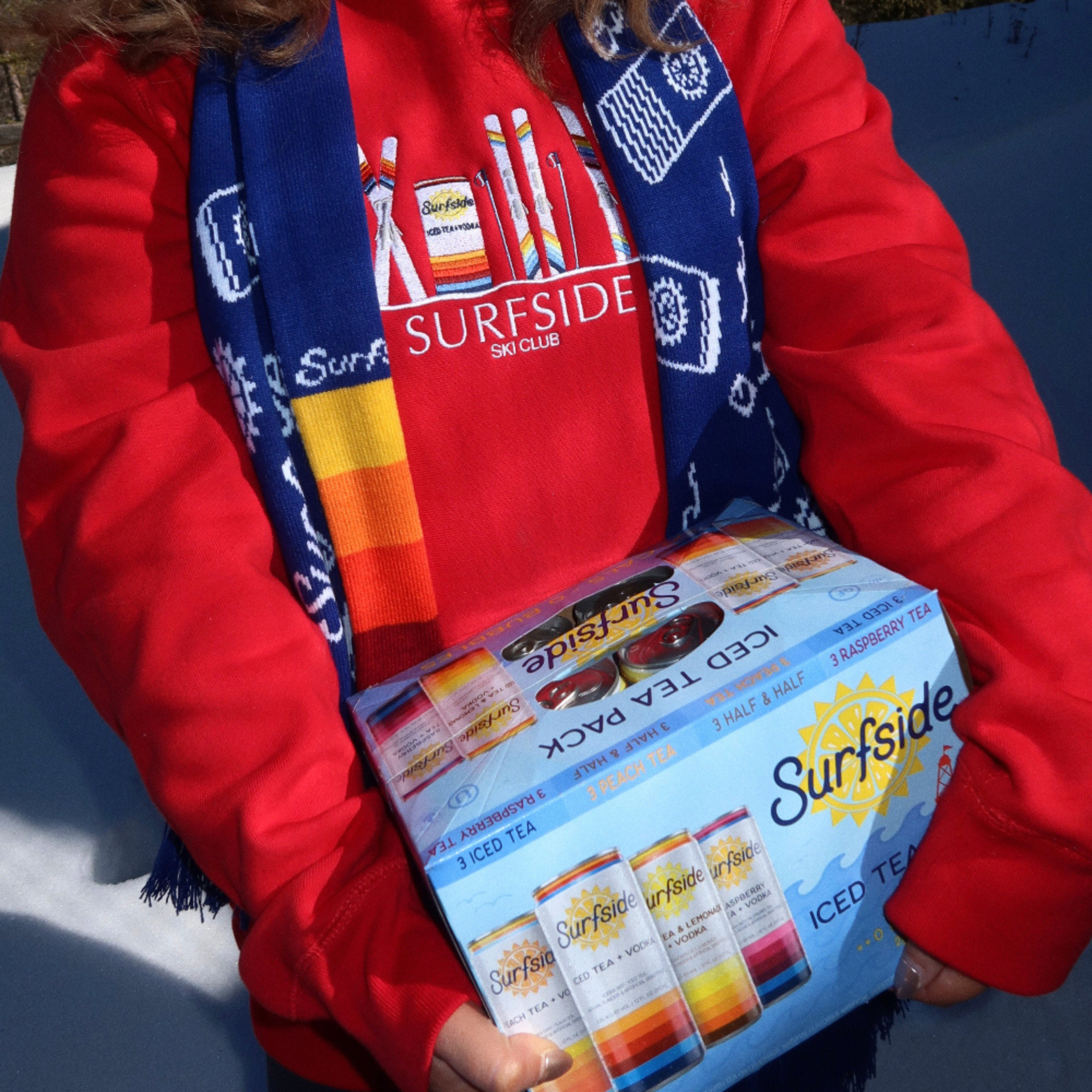 Person wearing a red jacket and blue scarf with 'Surfside' branding, holding a box of Surfside iced tea packs.