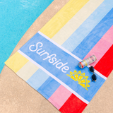 A colorful striped beach towel laid poolside featuring the text 'Surfside' and a yellow sun graphic. A can of Strawberry Lemonade and Vodka and a pair of sunglasses rest on the towel.