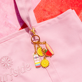 A gold-tone keychain charm set with colorful enamel pendants, including a surfboard, lemon slice, strawberry, sun, and striped Surfside strawberry lemonade and vodka can, attached to a pink tote bag with 'Surfside' embroidered on it.