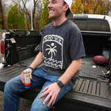 Man sitting in the back of a pickup truck holding a can of Surfside, with a 'Surfside Watering Hole' t-shirt on.