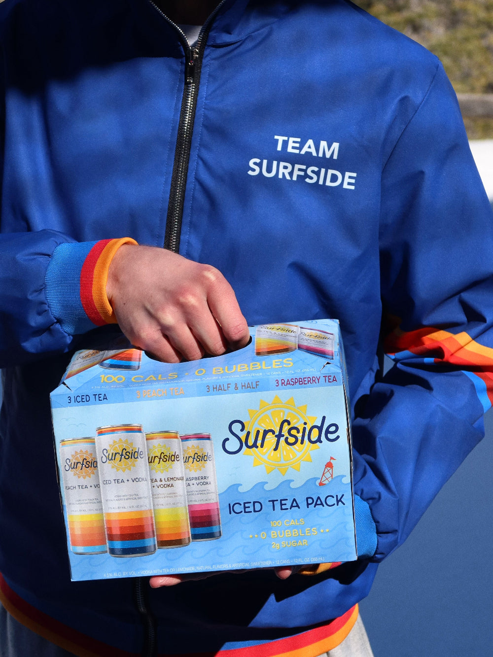Person wearing a blue Surfside windbreaker jacket with 'Team Surfrider' holding the Surfside Ice Tea Variety Pack