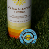 Surfside and Palm Golf Collaboration Ball marker resting in the grass on a Ice Tea and Lemonade and Vodka Surfside branded can