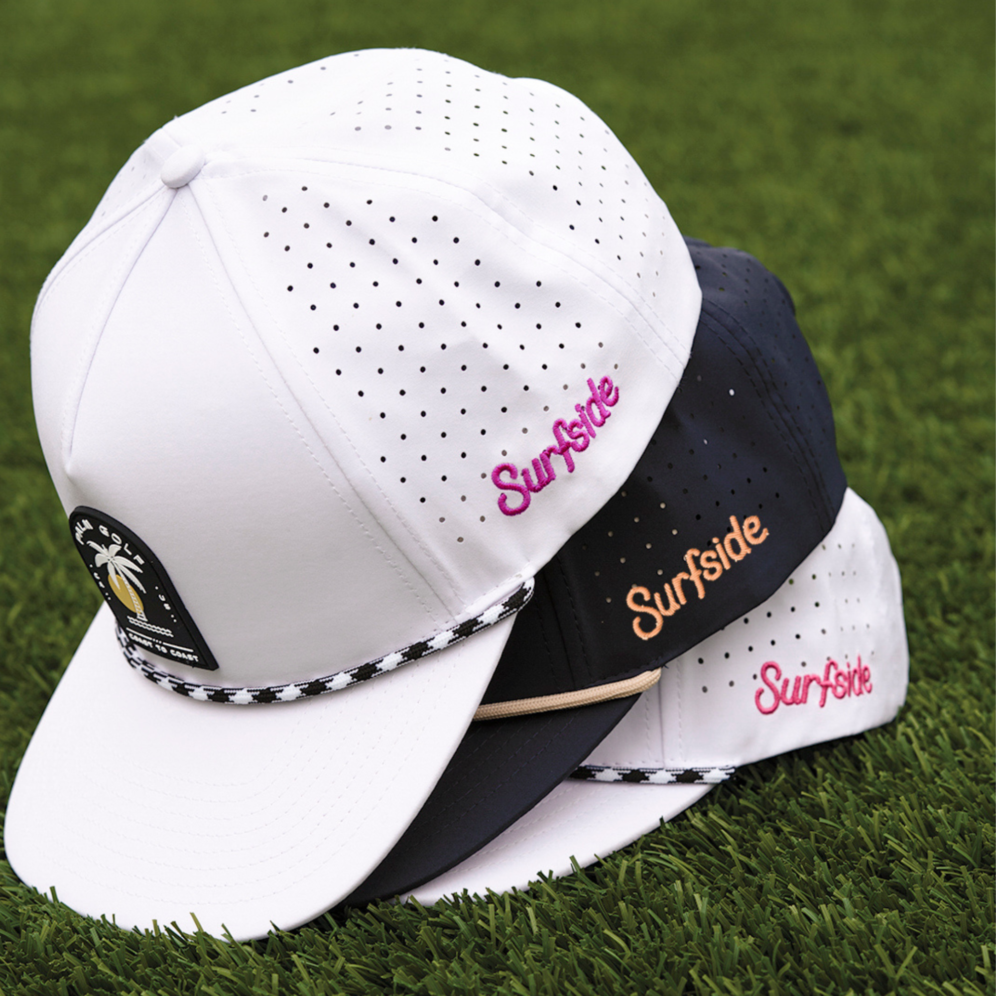Three stacked breathable Palm golf and Surfside collaborated hats. All have roped brimmed featuring the Surfside logo. Pictured on a grass background. 