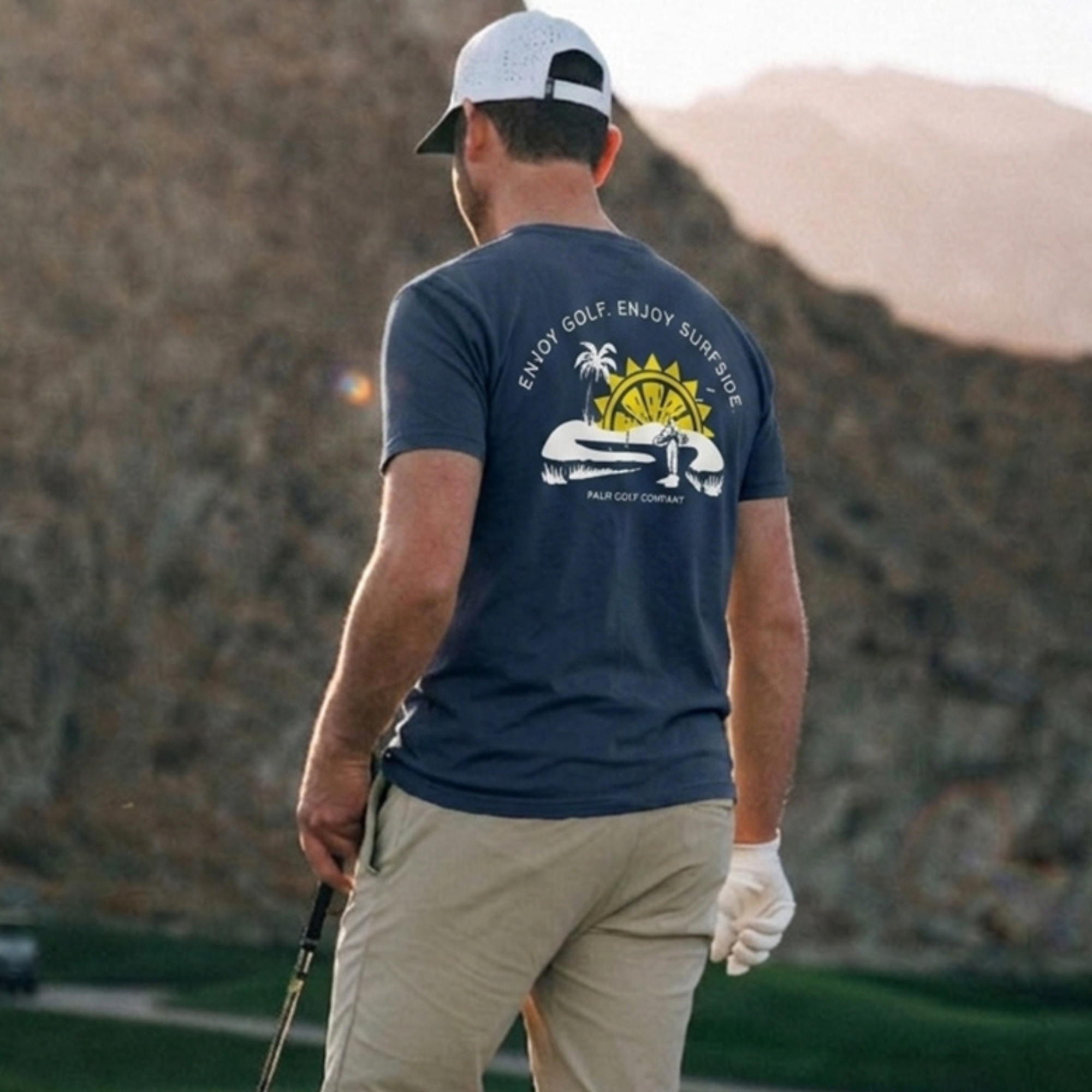 Back view of a man in a scenic background wearing a navy blue Surfside and Palm Golf T-shirt design featuring a golf course, sun, palm tree and 