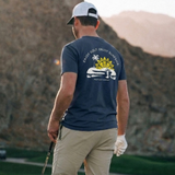 Back view of a man in a scenic background wearing a navy blue Surfside and Palm Golf T-shirt design featuring a golf course, sun, palm tree and "Enjoy Golf. Enjoy Surfside" text