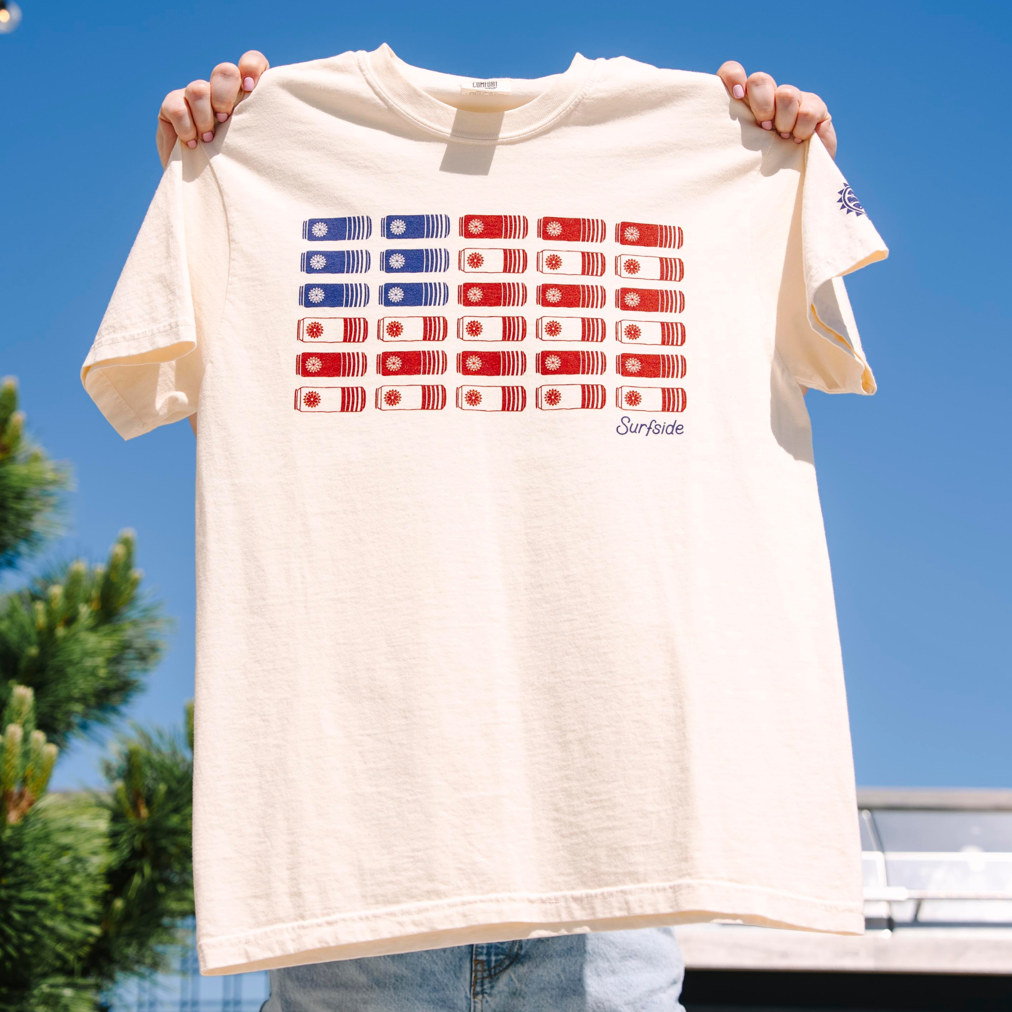 Beige short sleeve comfort colors t-shirt with red and blue Surfside cans in the form of an American Flag being held up against a clear blue sky. Surfside logo on the bottom left of the t-shirt and the surfside sun on bottom of the left sleeve