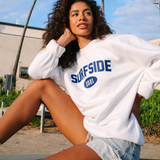 Woman wearing a white 'Surfside 2021' sweatshirt with palm trees and a building in the background.