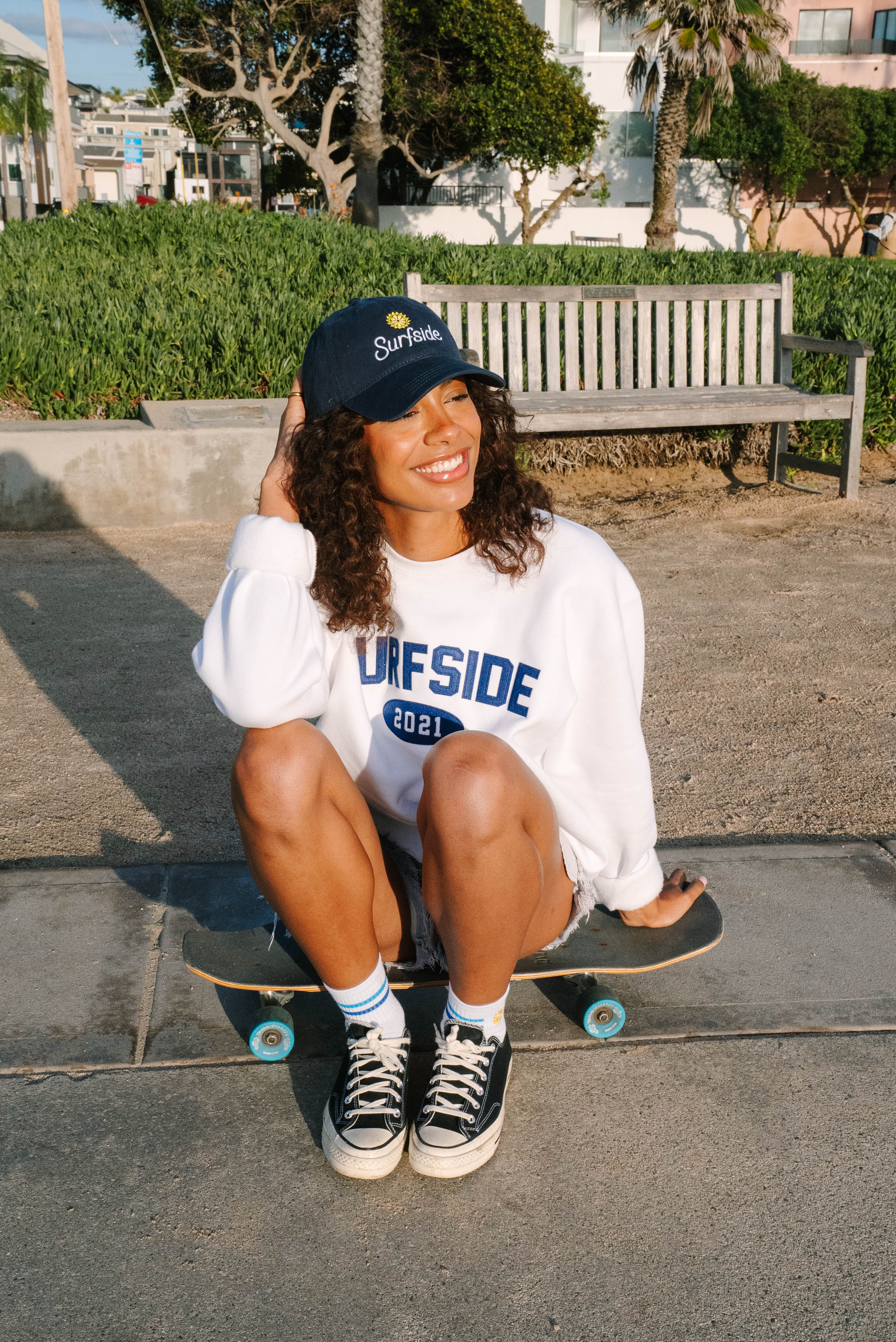 Person sitting on a skateboard wearing a white Surfside 2021 crewneck and a navy Surfside hat in an urban setting.