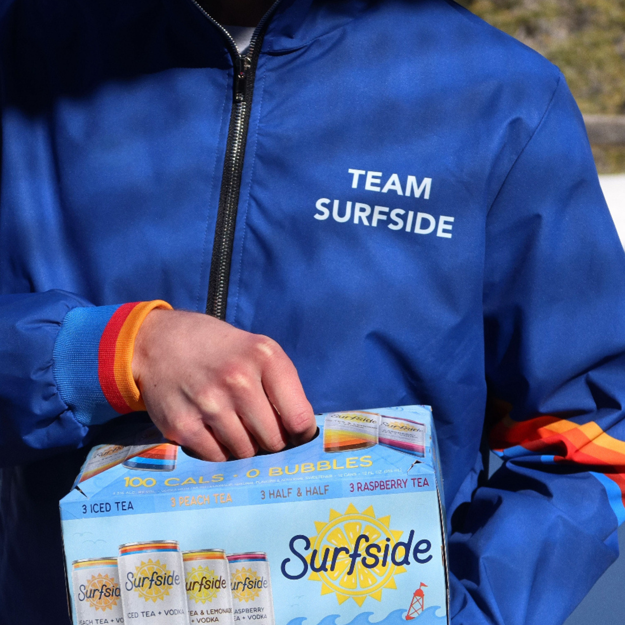 Person wearing a blue Surfside windbreaker jacket with 'Team Surfrider' holding the Surfside Ice Tea Variety Pack